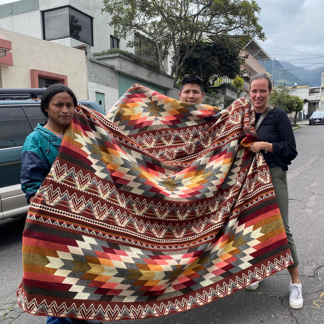 Drei Personen halten die große Alpaka-Decke 'Antisana Orange' im Freien hoch, um das vollständige Muster und die Größe zu zeigen.