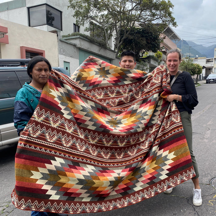 Drei Personen halten die große Alpaka-Decke 'Antisana Orange' im Freien hoch, um das vollständige Muster und die Größe zu zeigen.