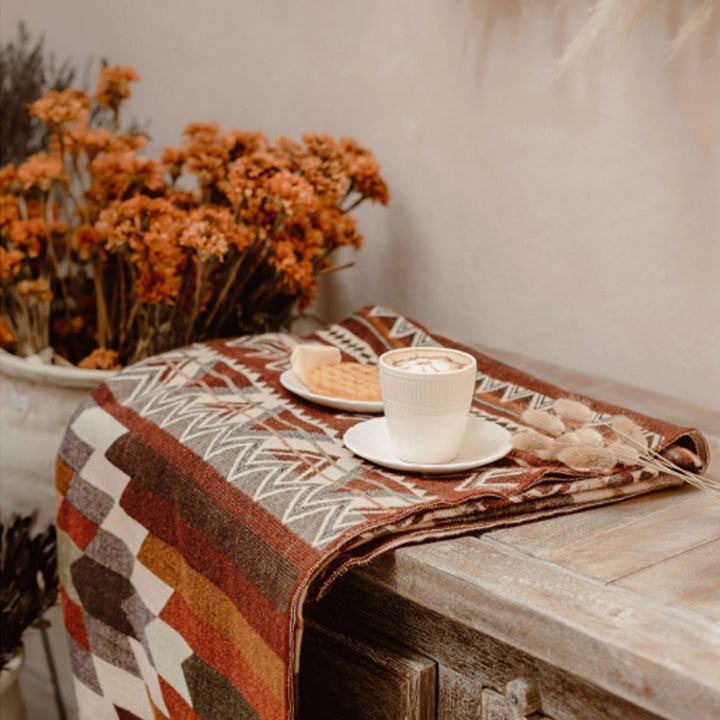 Nahaufnahme der gefalteten Alpaka-Decke 'Antisana Orange' auf einer Holzkommode mit einer Tasse Kaffee und getrockneten orangefarbenen Blumen in der Nähe.