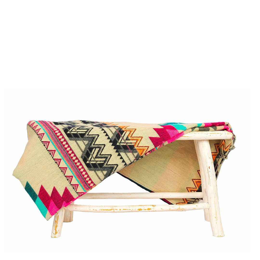 Alpaka-Decke 'Antisana Lila' über einem rustikalen weißen Hocker drapiert, zeigt die helle beige Seite mit farbigen Zickzack-Mustern.