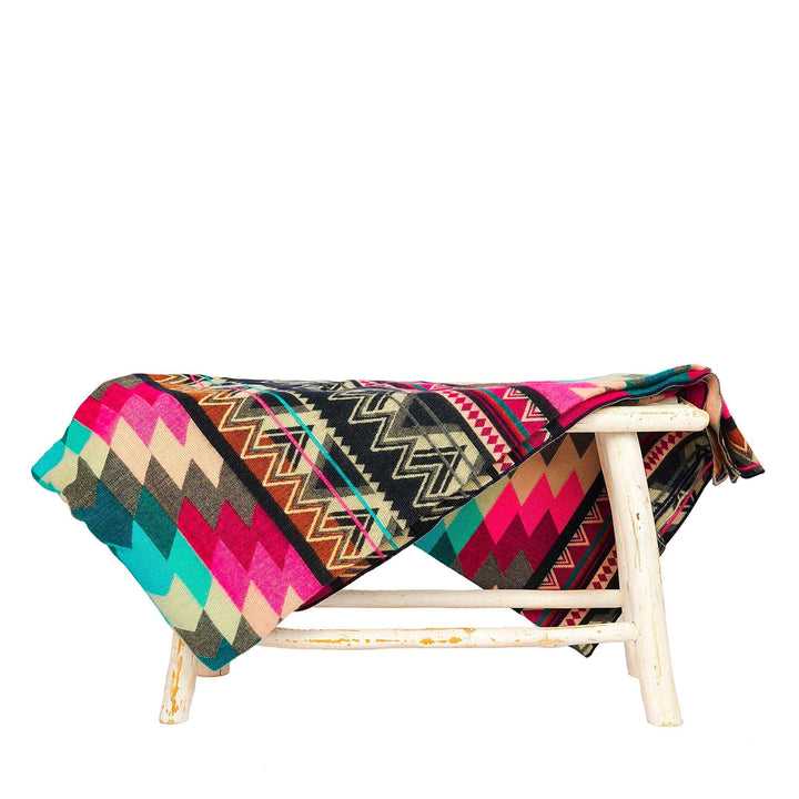 Alpaka-Decke 'Antisana Lila' über einem weißen Hocker, präsentiert die dunkle Seite mit kräftigen Pink-, Türkis- und Schwarz-Mustern.