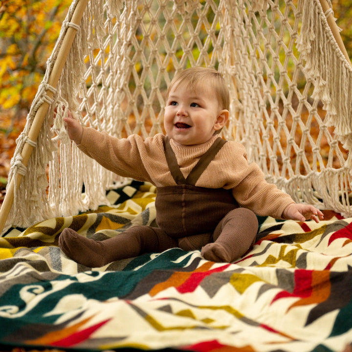 Glückliches Baby sitzt auf der bunten Alpaka-Decke 'Cayambe Grün' in einer Makramee-Schaukel im Herbstwald.