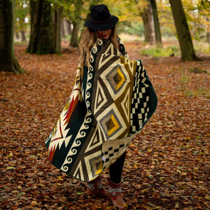 Frau im Herbstwald, eingewickelt in die große Alpaka-Decke 'Cayambe Grün' mit markanten Rautenmustern.