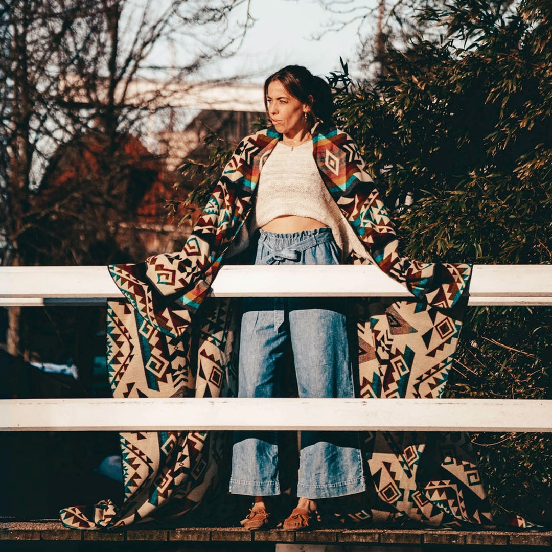 Frau steht im Freien und ist in die farbenfrohe Alpaka-Decke 'Chimborazo Mehrfarbig' gehüllt, die als Mantel dient.