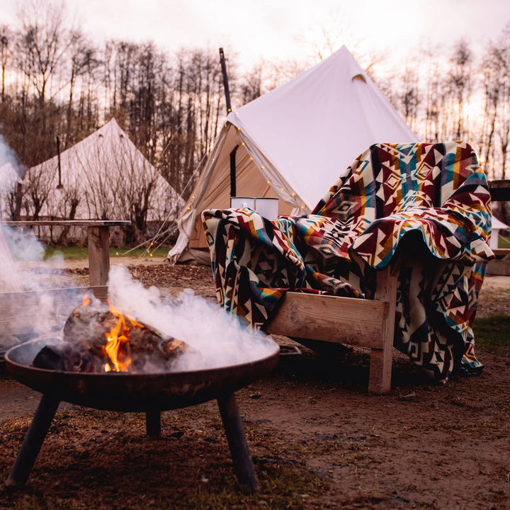 Gemütliche Glamping-Szene bei Sonnenuntergang mit der Alpaka-Decke 'Chimborazo Mehrfarbig' auf einem Holzstuhl und einer Feuerschale im Vordergrund.