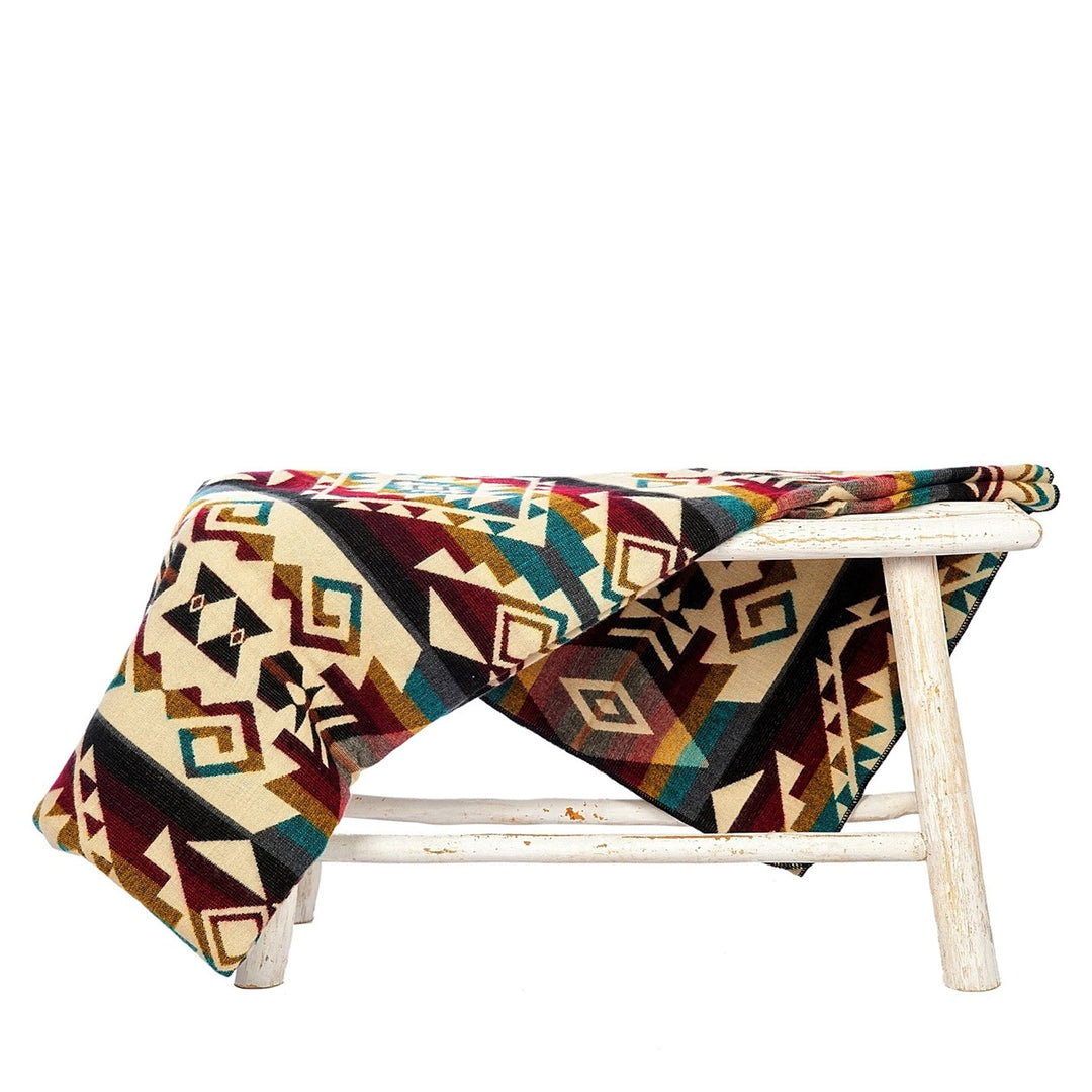 Alpaka-Decke 'Chimborazo Mehrfarbig' über einem rustikalen weißen Hocker drapiert, zeigt das farbenfrohe, indigene Muster.