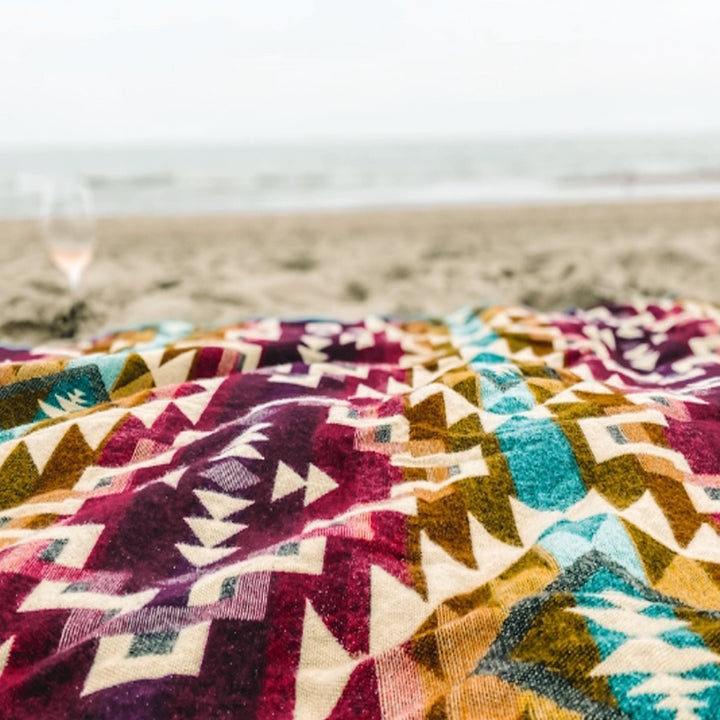 Nahaufnahme der farbenfrohen Alpaka-Decke 'Chimborazo Rosa' als Picknickdecke am Strand, im Hintergrund das Meer.