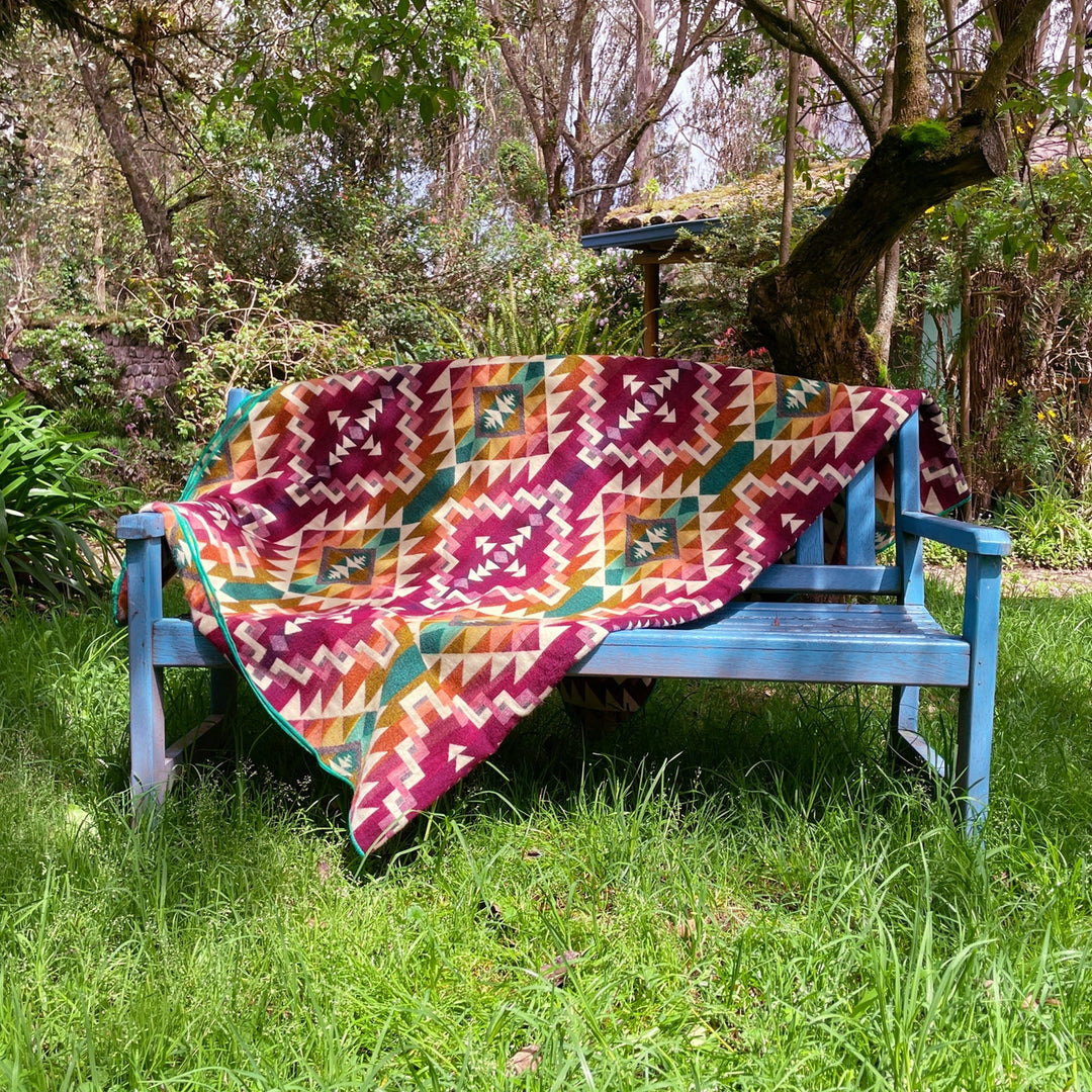 Alpaka-Decke 'Chimborazo Rosa' drapiert über einer blauen Gartenbank in einem grünen, sonnigen Garten.