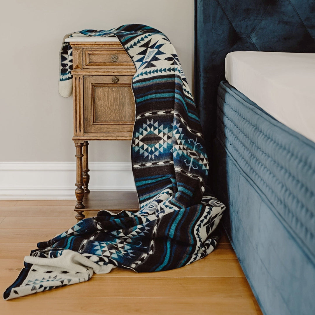 Alpaka-Decke 'Cotopaxi Blau' drapiert über einer antiken Nachtkommode neben einem blauen Polsterbett.
