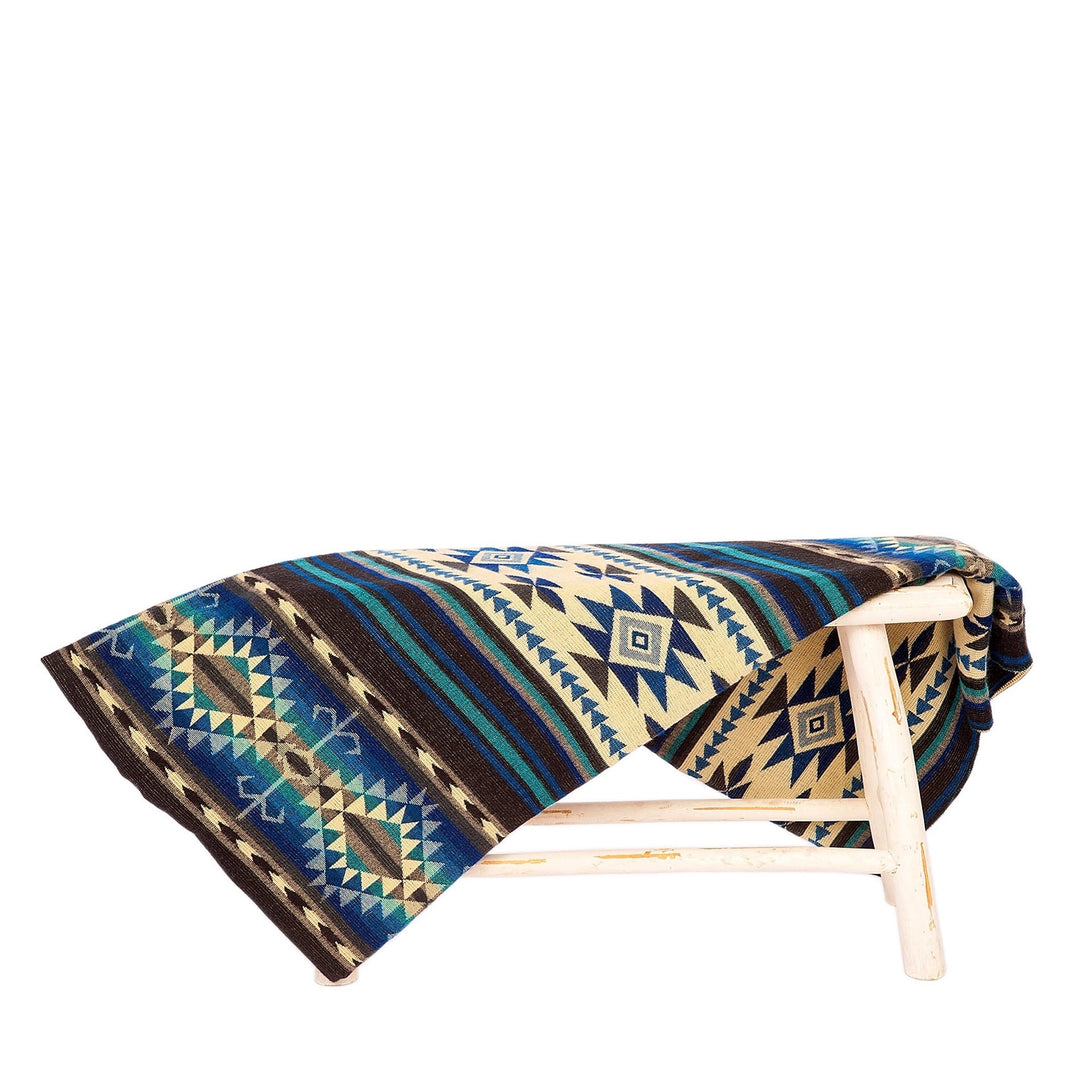 Alpaka-Decke 'Cotopaxi Blau' über einem rustikalen weißen Hocker drapiert, zeigt das blaue, beige und braune Muster.