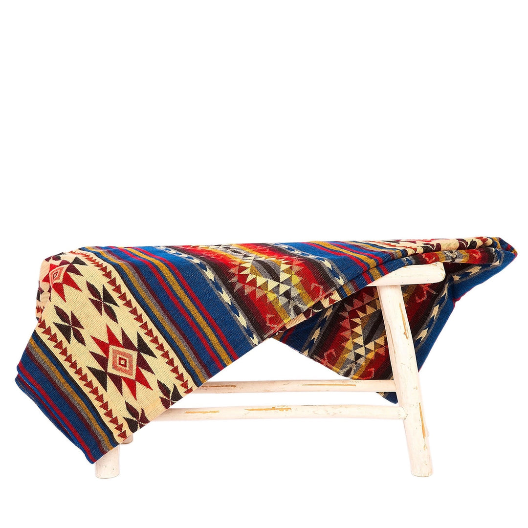 Alpaka-Decke 'Cotopaxi Mehrfarbig' über einem rustikalen weißen Hocker drapiert, zeigt die farbenfrohen Streifen und Stammesmuster.