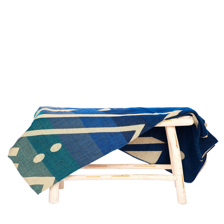 Alpaka-Decke 'Imbabura Blau' über einem rustikalen weißen Hocker drapiert, betont die Blautöne und das indigene Muster.