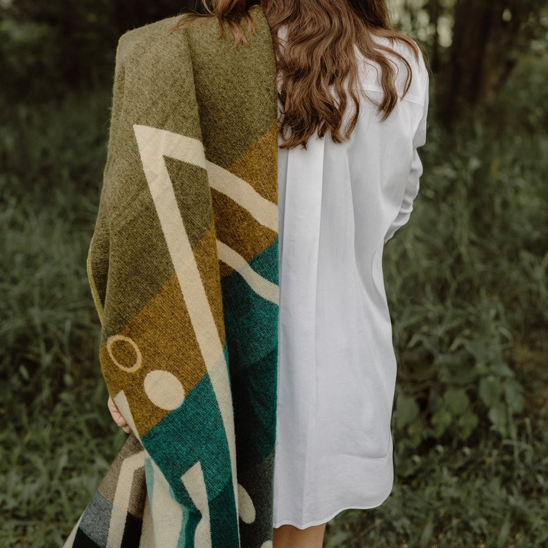 Rückenansicht einer Person mit langen, gewellten braunen Haaren, die ein weißes Hemd trägt und eine Alpaka-Decke mit einem großflächigen geometrischen Muster in Oliv, Gelb und Petrol über die Schulter hält. Die Szene spielt in einem Wald oder Park