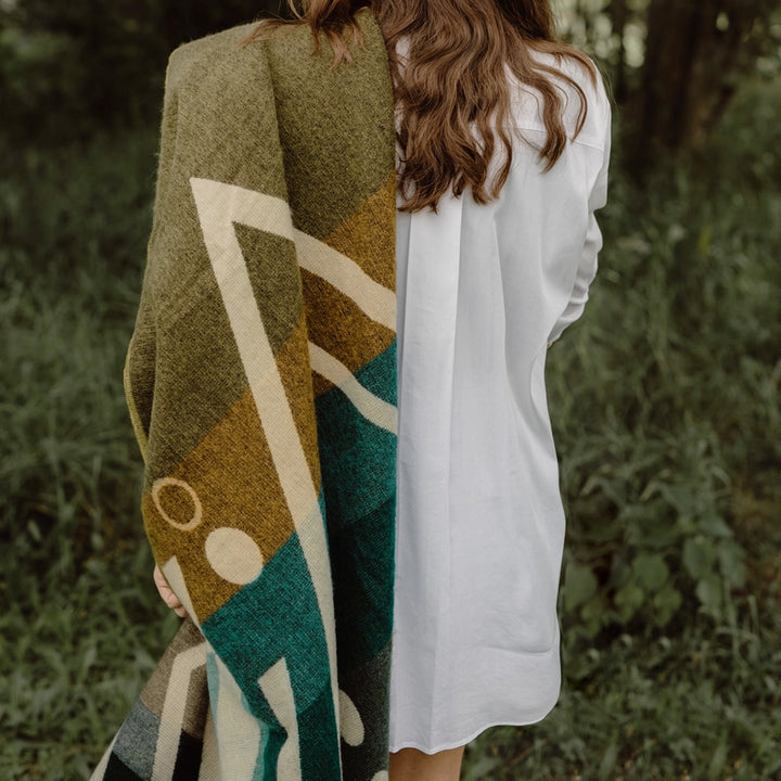 Rückenansicht einer Person mit langen, gewellten braunen Haaren, die ein weißes Hemd trägt und eine Alpaka-Decke mit einem großflächigen geometrischen Muster in Oliv, Gelb und Petrol über die Schulter hält. Die Szene spielt in einem Wald oder Park