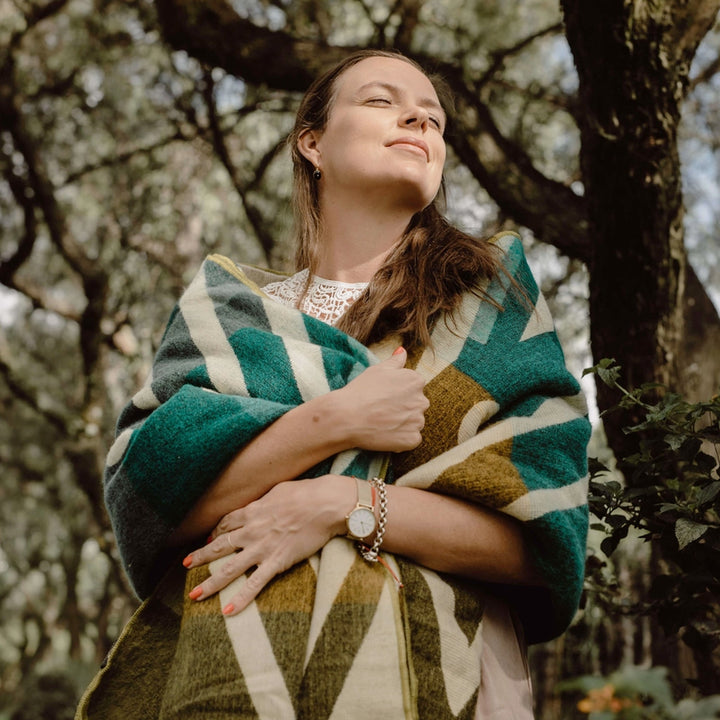 Eine Person mittleren Alters, die eine Alpaka-Decke mit breiten, geometrischen Streifen in Oliv, Beige und Türkis um die Schultern geschlungen hat. Sie steht in einem Waldstück und blickt mit geschlossenen Augen leicht nach oben, das Gesicht von der Sonne beschienen