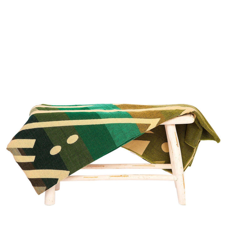 Eine Alpaka-Decke mit einem großflächigen geometrischen Muster in Grüntönen, Beige und Oliv liegt über einer kleinen, rustikalen weißen Holzbank. Der Hintergrund ist weiß
