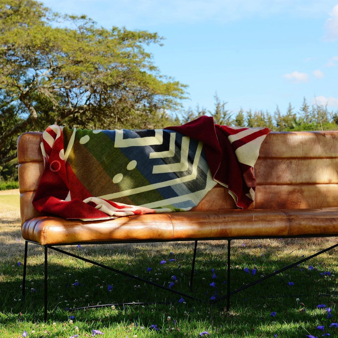 Eine Alpaka-Decke mit einem großflächigen, bunten geometrischen Muster liegt auf einer modernen, braunen Lederbank mit einem schwarzen Metallgestell. Die Szene ist im Freien auf einer grünen Wiese mit violetten Blumen im Vordergrund und Bäumen im Hintergrund unter blauem Himmel