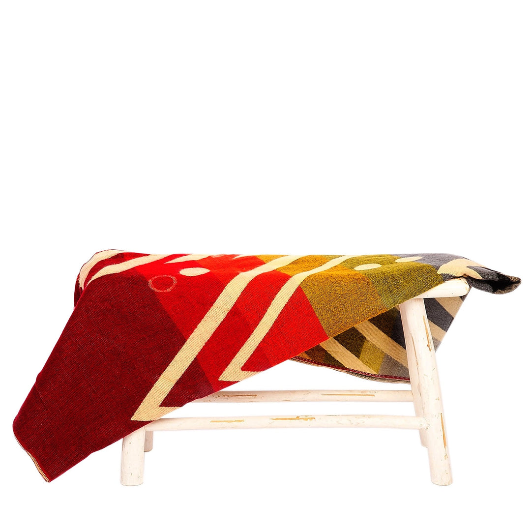 Eine Alpaka-Decke mit einem großflächigen geometrischen Muster in Rot, Gelb, Oliv und Blau liegt über einer kleinen, rustikalen weißen Holzbank. Der Hintergrund ist weiß