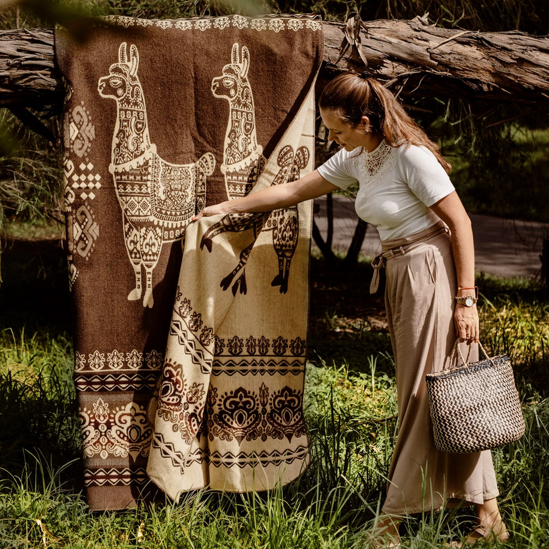 Eine Frau in einem weißen Oberteil und beigen weiten Hosen steht in einer natürlichen Umgebung (Wiese, Bäume) und berührt eine große, braune Alpaka-Decke mit weißen Lama-Motiven, die an einem Baumstamm hängt. Sie hält eine gewebte Tasche in ihrer linken Hand