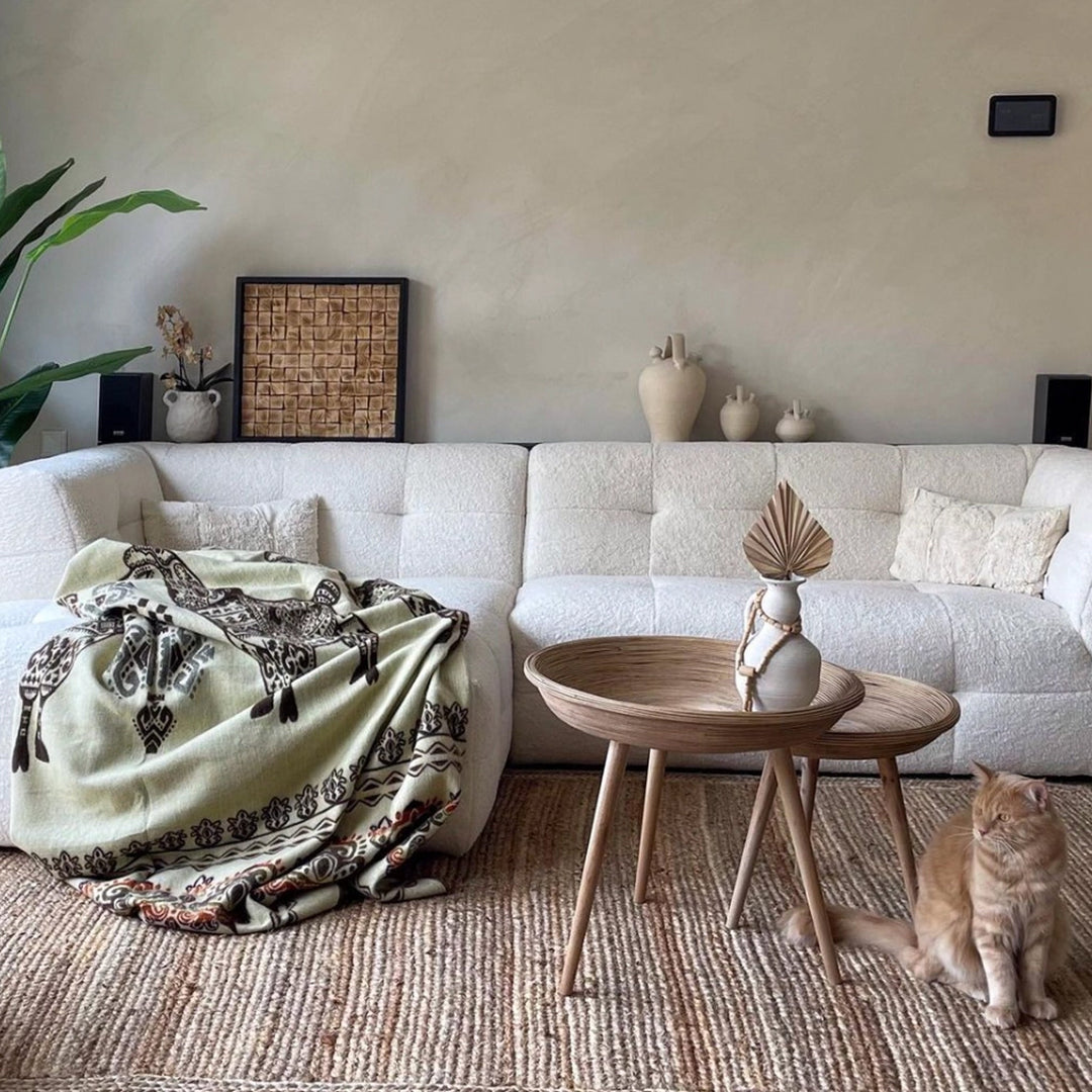 Ein modernes Wohnzimmer mit einem beigefarbenen, strukturierten Sofa und zwei runden Holztischen. Auf dem Sofa liegt eine helle Alpaka-Decke mit Lama-Motiven in Braun. Ein ingwerfarbenes (orange-braunes) Kätzchen sitzt rechts unten auf einem beigen Jute-Teppich. Dekorative Vasen und Pflanzen ergänzen das Bild