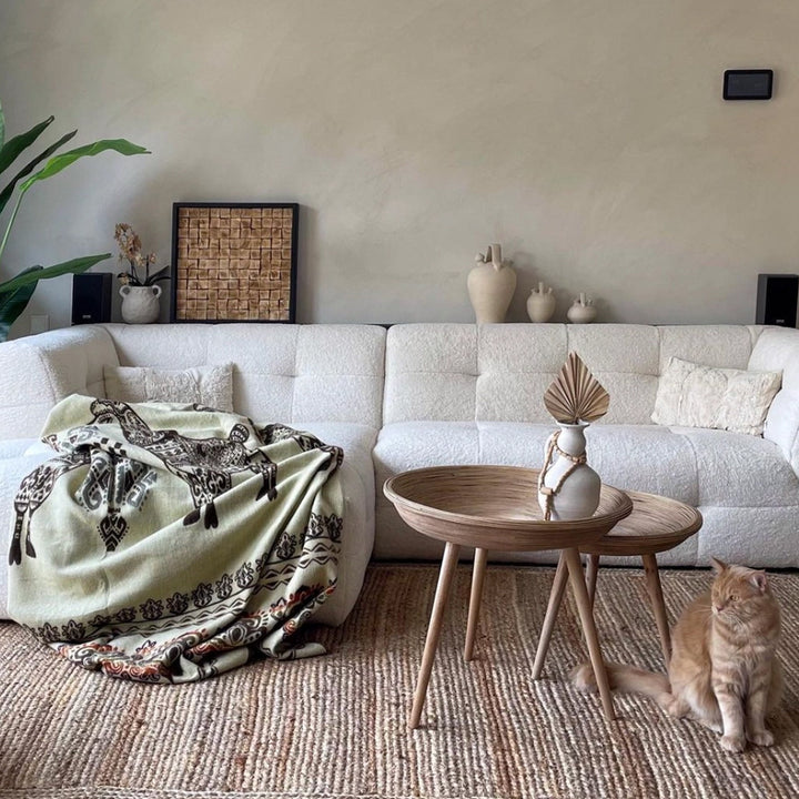 Ein modernes Wohnzimmer mit einem beigefarbenen, strukturierten Sofa und zwei runden Holztischen. Auf dem Sofa liegt eine helle Alpaka-Decke mit Lama-Motiven in Braun. Ein ingwerfarbenes (orange-braunes) Kätzchen sitzt rechts unten auf einem beigen Jute-Teppich. Dekorative Vasen und Pflanzen ergänzen das Bild
