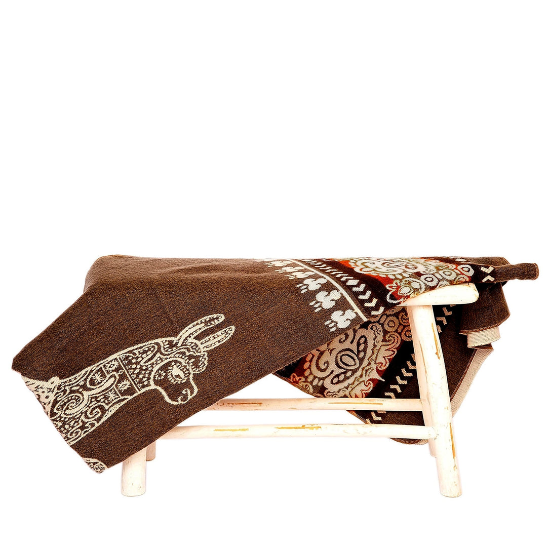 Eine dunkelbraune Alpaka-Decke mit einem weißen Lama-Kopfmotiv an einer Ecke liegt über einer kleinen, rustikalen weißen Holzbank. Die Decke ist teilweise umgeschlagen und zeigt auf der Rückseite ein helleres, florales Muster. Der Hintergrund ist weiß