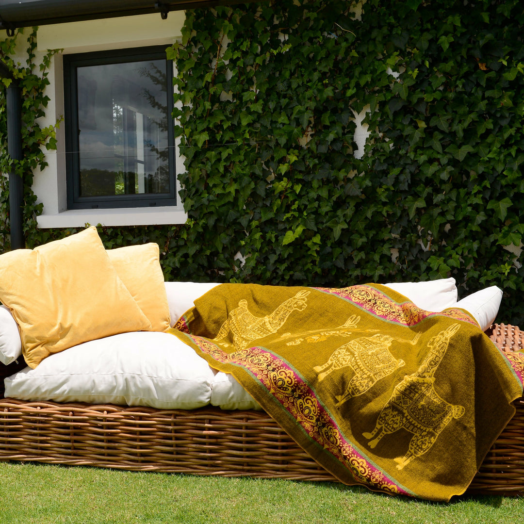 Eine Alpaka-Decke in Senfgelb mit gelben Lama-Motiven und fuchsiafarbenen Bordüren liegt auf einem weißen Polster eines Korbmöbel-Sofas im Freien. Auf dem Sofa liegen auch zwei gelbe Kissen. Das Sofa steht auf einer grünen Wiese vor einer weißen Hauswand, die größtenteils mit Efeu bewachsen ist