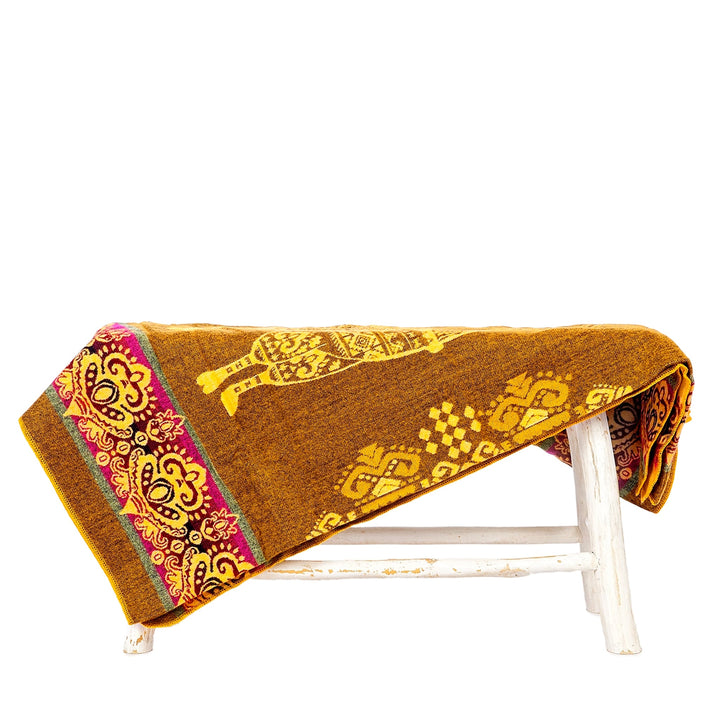 Eine senfgelbe Alpaka-Decke mit einem gelben Lama-Motiv und einer fuchsiafarbenen Bordüre liegt über einer kleinen, rustikalen weißen Holzbank. Die Decke ist teilweise umgeschlagen. Der Hintergrund ist weiß
