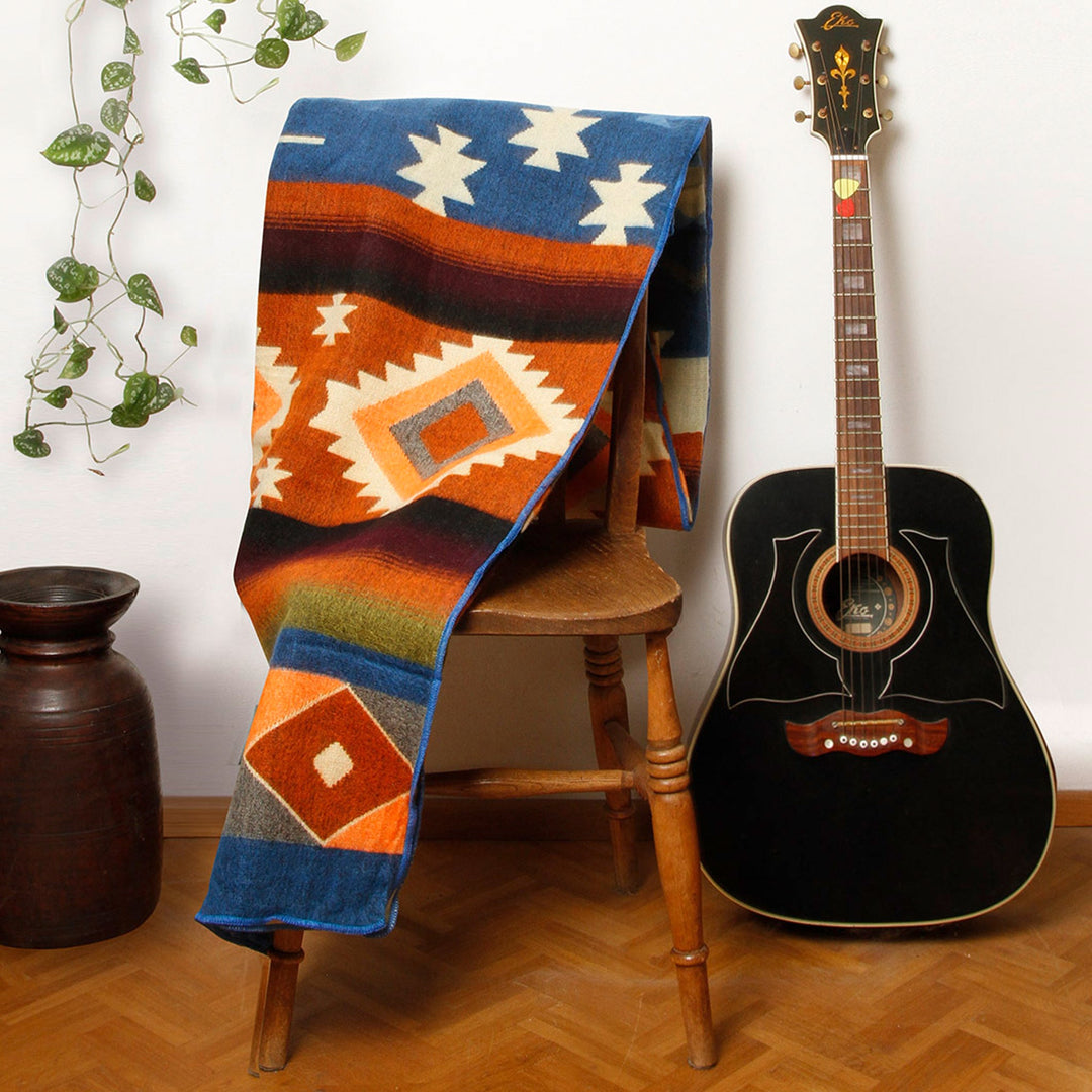 Eine Alpaka-Decke mit dem bunten geometrischen Muster liegt über einer hölzernen Stuhllehne. Rechts daneben steht eine schwarze Akustikgitarre, links ein brauner Tonkrug und eine Hängepflanze. Die Szene ist in einem Innenraum mit Holzboden