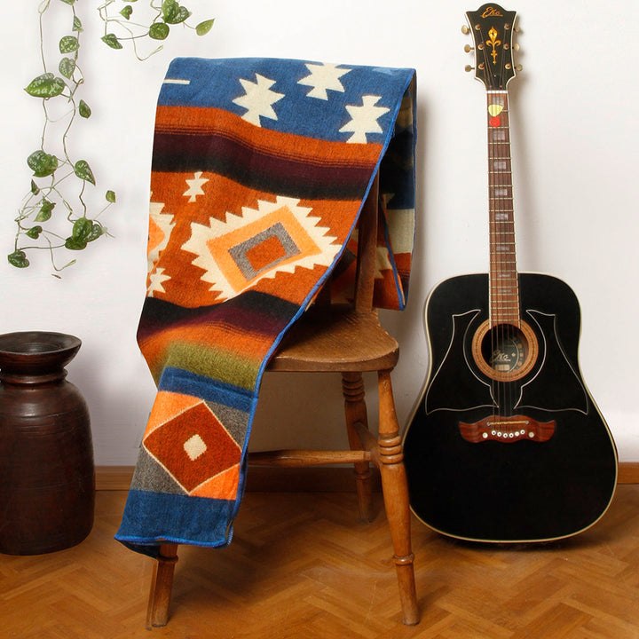 Eine Alpaka-Decke mit dem bunten geometrischen Muster liegt über einer hölzernen Stuhllehne. Rechts daneben steht eine schwarze Akustikgitarre, links ein brauner Tonkrug und eine Hängepflanze. Die Szene ist in einem Innenraum mit Holzboden