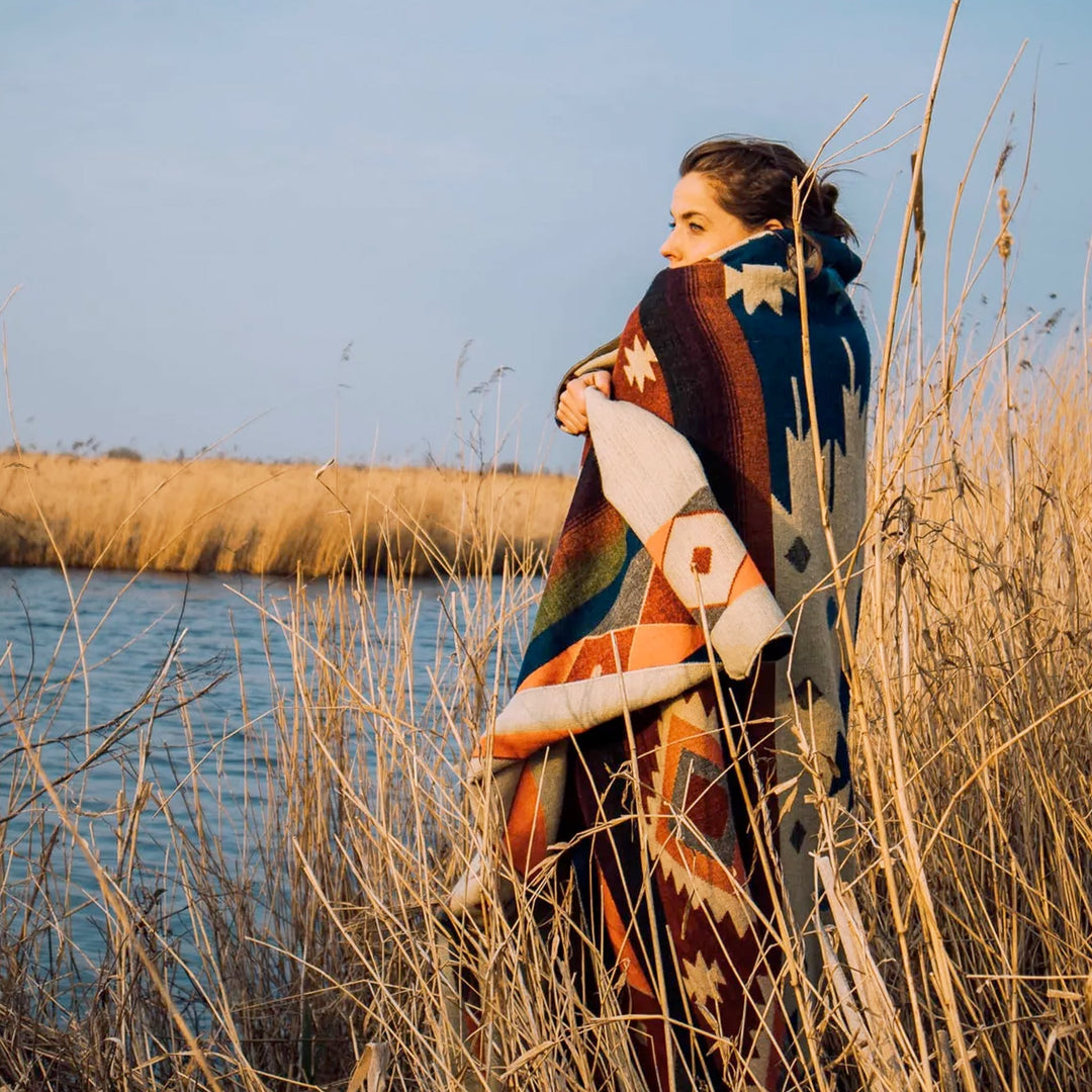 Eine Person ist in eine bunte Alpaka-Decke mit geometrischen Mustern gehüllt und steht in hohem, trockenem Schilf oder Gras am Ufer eines Gewässers. Die Abendsonne beleuchtet die Szene