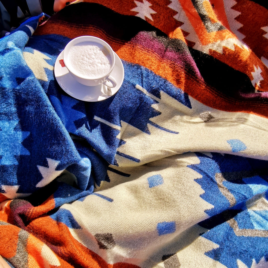 Nahaufnahme einer weißen Tasse mit Milchschaum auf einer weißen Untertasse, die auf der bunten Alpaka-Decke liegt. Die Decke ist zerknittert und zeigt die geometrischen Muster in leuchtendem Blau, Orange, Rot und Beige im Sonnenlicht
