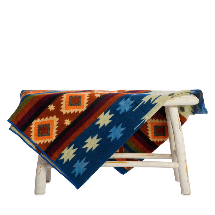 Die Alpaka-Decke mit dem bunten geometrischen Muster liegt über einer kleinen, rustikalen weißen Holzbank. Der Hintergrund ist weiß