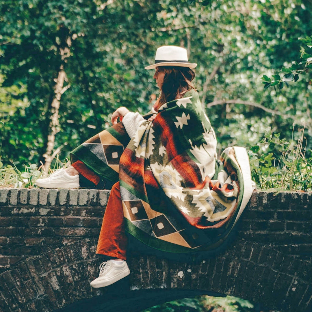 Eine Frau mit einem weißen Hut und orangeroter Hose sitzt auf einer alten Stein- oder Ziegelmauer in einem Waldgebiet. Sie ist in eine Alpaka-Decke mit einem bunten geometrischen Muster in Grün, Orange und Beige eingehüllt