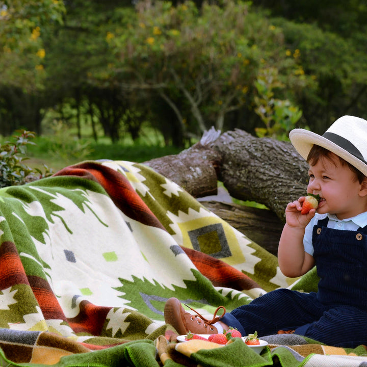 Ein Kleinkind in dunkelblauer Latzhose und einem weißen Hut sitzt im Freien auf einer Alpaka-Decke mit grünem, orangem und beigem geometrischem Muster und isst eine Erdbeere. Mehrere Erdbeeren liegen auf einem kleinen Teller neben dem Kind. Die Szene ist in einer grünen, natürlichen Umgebung