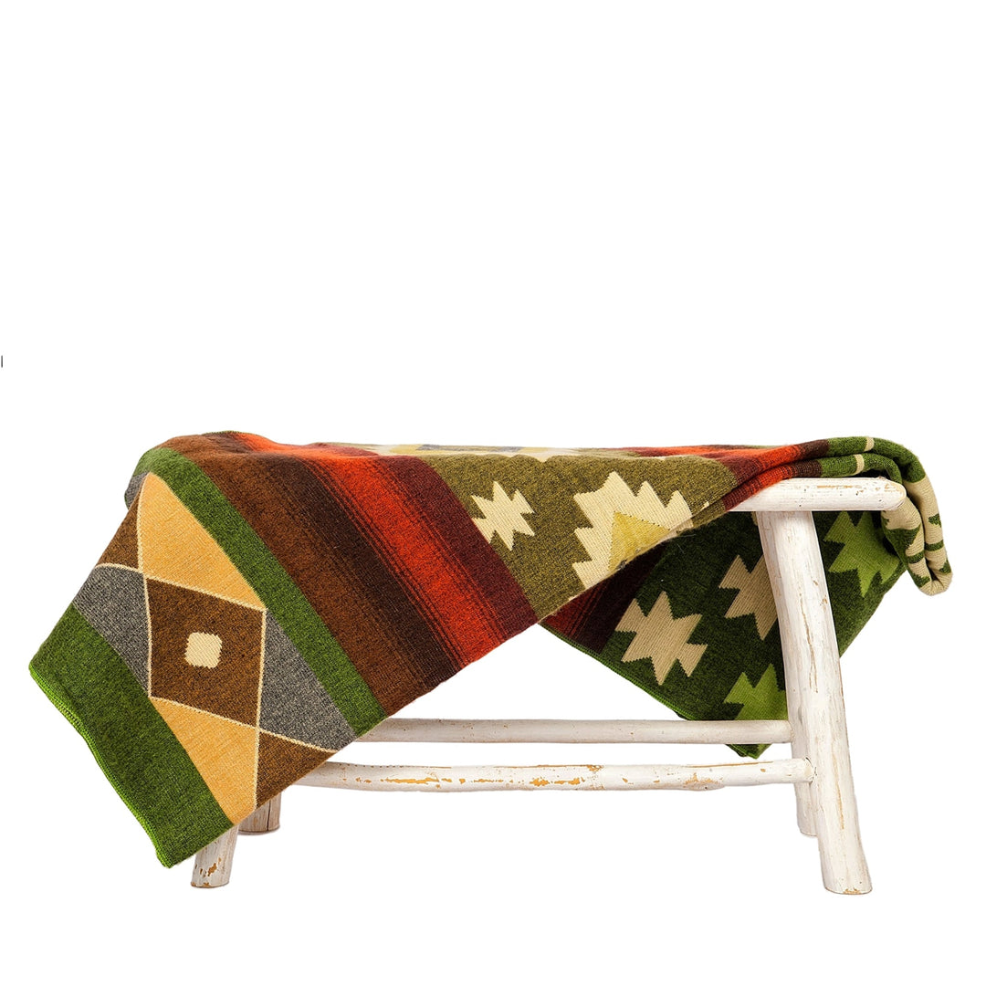 Die Alpaka-Decke mit dem bunten geometrischen Muster in Grün, Orange, Braun und Gelb liegt über einer kleinen, rustikalen weißen Holzbank. Der Hintergrund ist weiß