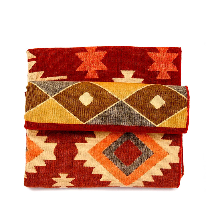 Die Alpaka-Decke mit dem bunten geometrischen Muster in Rot, Beige, Orange, Braun und Gelb ist dreifach gefaltet und gestapelt dargestellt, wobei verschiedene Musterabschnitte sichtbar sind. Der Hintergrund ist weiß