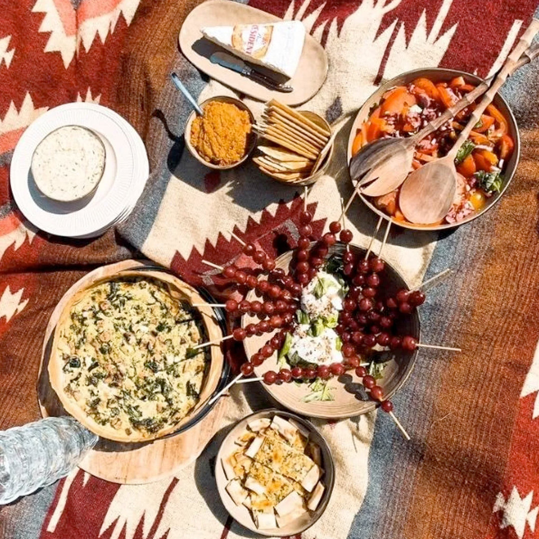Eine Decke mit rotem und beigem geometrischem Muster dient als Unterlage für ein Picknick. Darauf stehen verschiedene Speisen in Schüsseln und auf Tellern, darunter eine Quiche, ein Salat, Käse und Traubenspieße. Die Aufnahme ist von oben