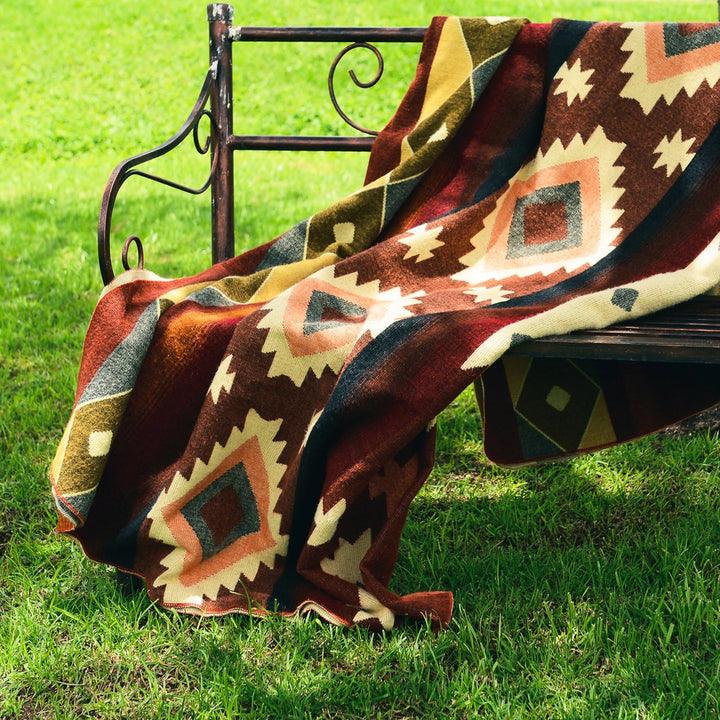 Eine Alpaka-Decke mit einem großflächigen, rot-braunen und gelben geometrischen Muster liegt lässig auf einer dunklen, schmiedeeisernen Gartenbank. Die Bank steht auf einer grünen Rasenfläche