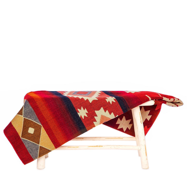 Die Alpaka-Decke mit dem bunten geometrischen Muster in Rot, Orange, Blau und Erdtönen liegt über einer kleinen, rustikalen weißen Holzbank. Der Hintergrund ist weiß