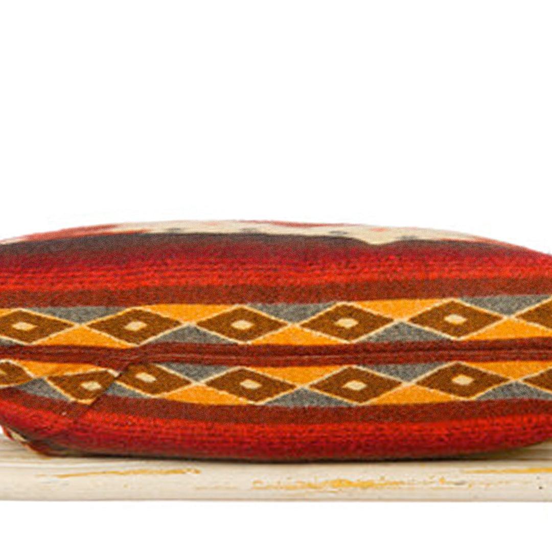 Nahaufnahme der Seite eines rechteckigen Alpaka-Kissens, das ein Muster aus horizontalen Streifen und einer Reihe kleiner Rauten in Orange, Rot, Braun und Grau zeigt. Das Kissen liegt auf einer hellen, rustikalen Oberfläche