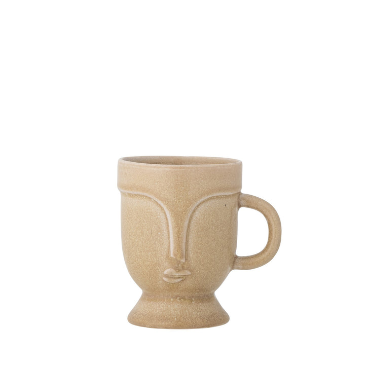 beige Keramiktasse mit Henkel, kleinem Standfuß und stilisiertem Gesichtsrelief auf der Vorderseite