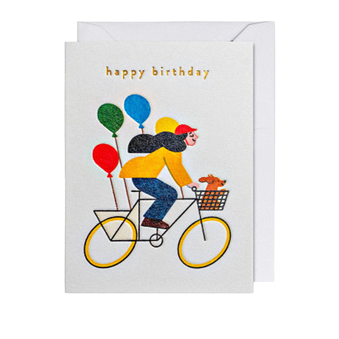 Geburtstagskarte mit „happy birthday“-Schriftzug, Frau auf Fahrrad mit bunten Ballons und kleinem Hund im Fahrradkorb.