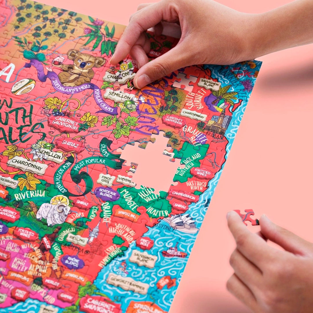 Nahaufnahme von Händen, die ein Puzzleteil in die Region New South Wales des "Australien Wein-Puzzles" einsetzen, mit Details zu Rebsorten wie Shiraz, Semillon und Chardonnay.