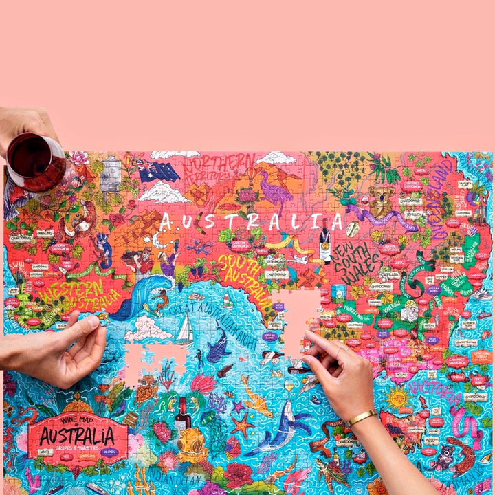 Zwei Hände beim Zusammenfügen des 1000-Teile-Puzzles der australischen Weinregionen, mit einem Glas Rotwein am oberen linken Rand.