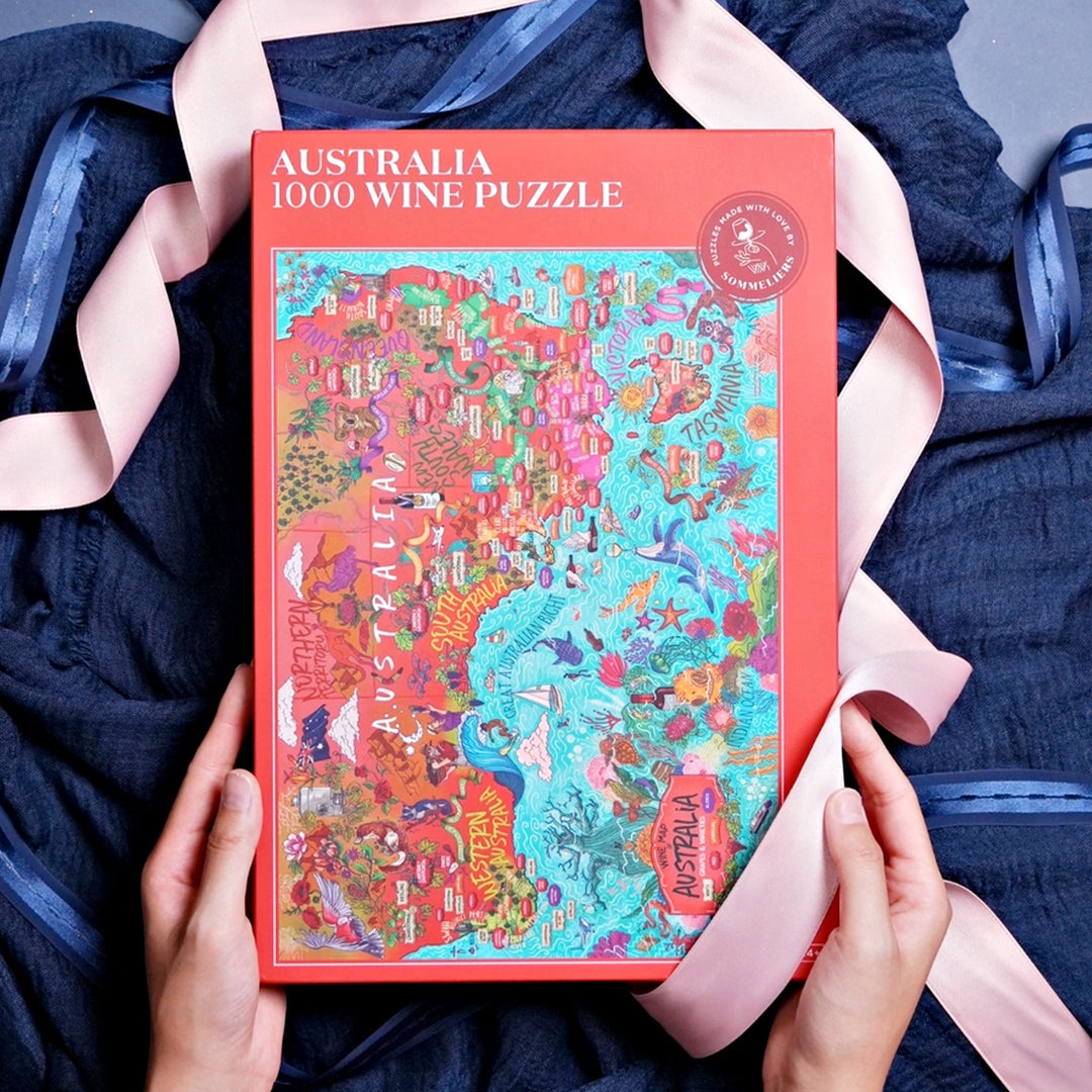 Geschlossene rote Box des "Australien 1000 Wein-Puzzles" mit der illustrierten Weinlandkarte, umrahmt von einem dunkelblauen Tuch und einem rosa Band, gehalten von zwei Händen.