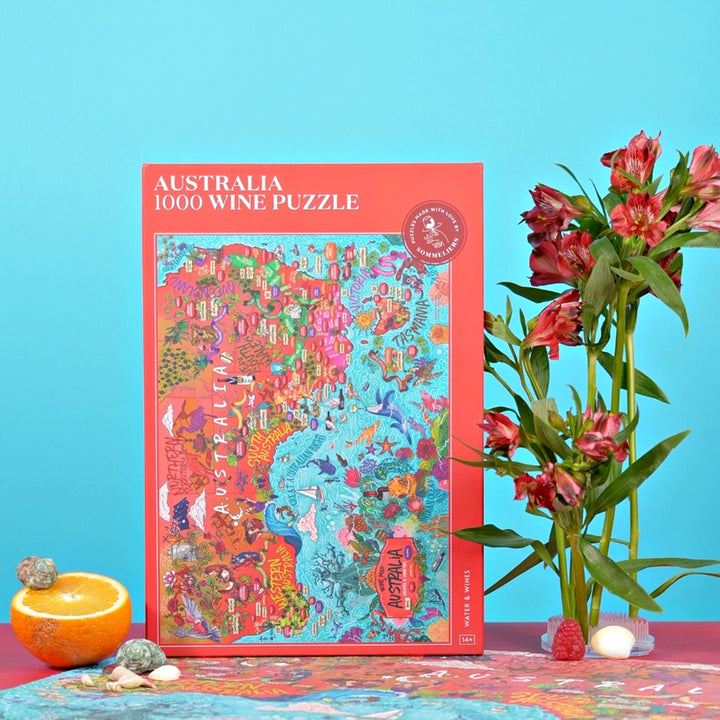 Produktaufnahme der roten Puzzle-Box "Australien 1000 Wein-Puzzle" mit einem Strauß roter Blumen, einer Orange und einer blauen Hintergrundwand.