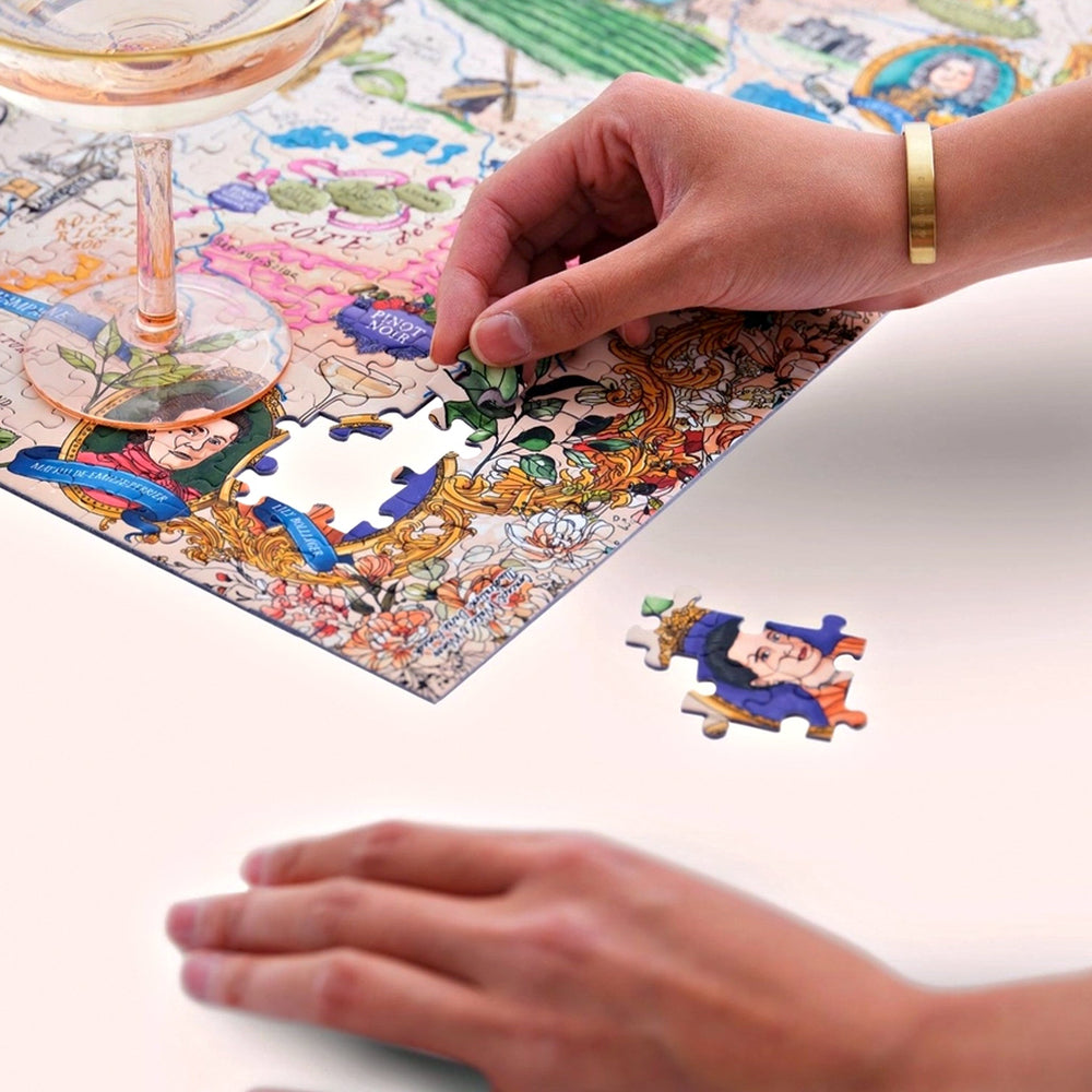 Nahaufnahme von Händen, die ein Puzzleteil mit einem historischen Porträt in das unvollendete Champagner-Puzzle einsetzen. Ein Sektglas ist im Vordergrund platziert.