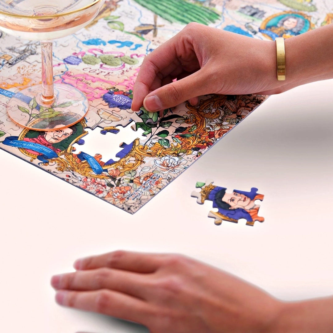 Nahaufnahme von Händen, die ein Puzzleteil mit einem historischen Porträt in das unvollendete Champagner-Puzzle einsetzen. Ein Sektglas ist im Vordergrund platziert.