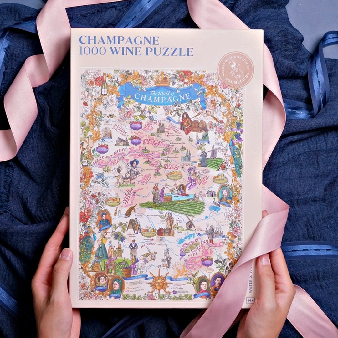 Geschlossene Puzzle-Box "Champagne 1000 Wine Puzzle", gehalten von Händen, umrahmt von einem dunkelblauen Tuch und einem rosa Satinband.